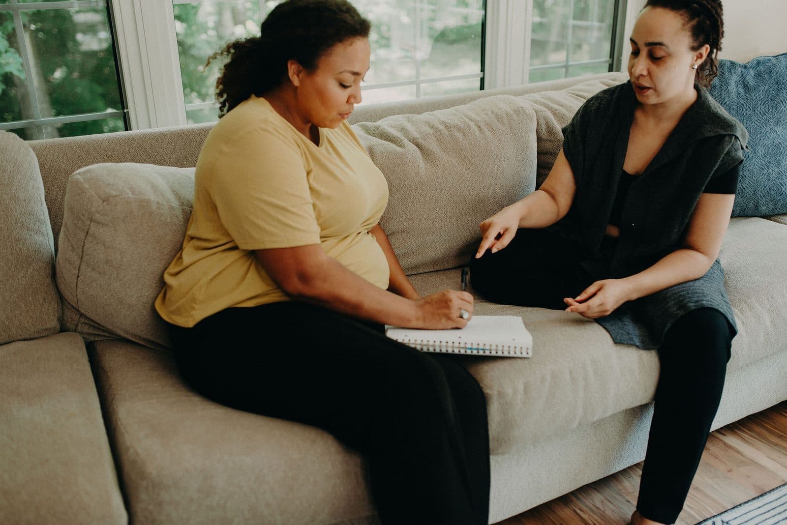 A doula sits with a pregnant client on a couch, writing on a notepad as they talk.